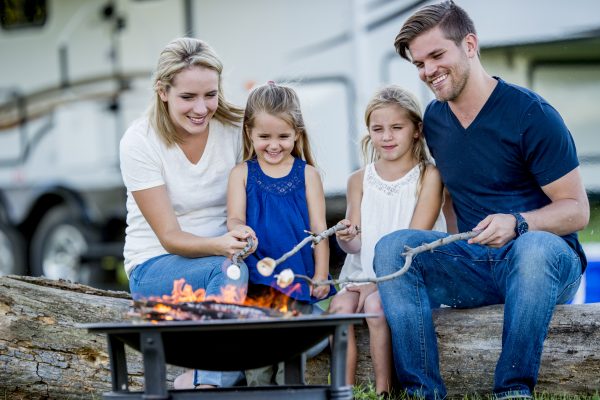 A Caucasian mother, father, and two daughters are camping together. Their trailer is in the background. They are sitting on logs and roasting marshmallows over a campfire.